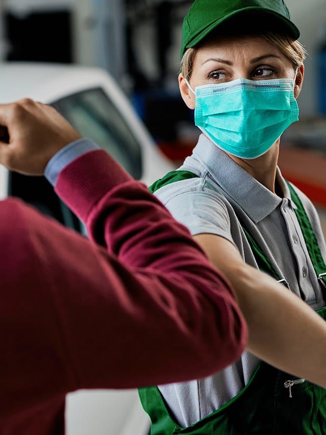 Avaliadora InstaCarro usando máscara comprimentando cliente com toque no cotovelo. Ao fundo um carro - instacarro chega a cotia